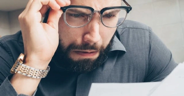 Man wearing glasses, hands on his forehead, reading a document with a worried expression.