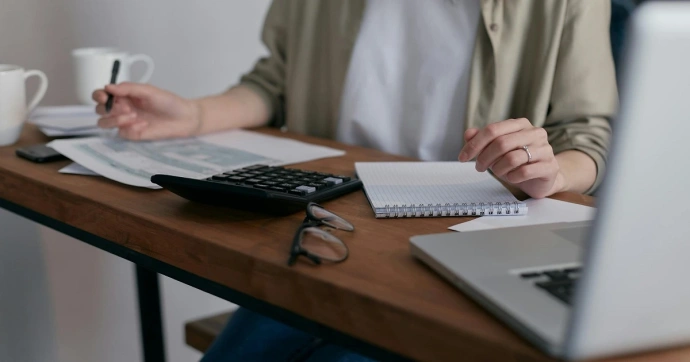 Person writing in a notebook, with a calculator and a laptop nearby, doing calculations.